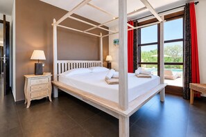 A four-poster bed with white linens, two bedside tables with lamps, and a view of a pool through large windows.