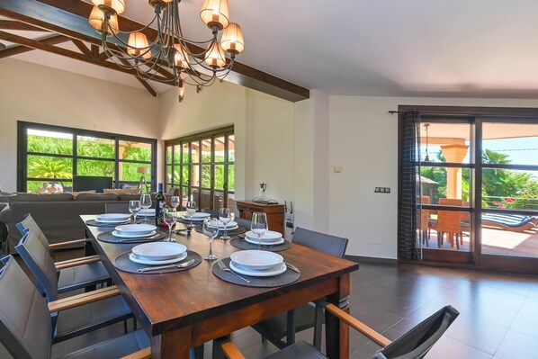 A dining area with a wooden table set for six, large windows, and a view of greenery outside.