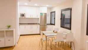 Una cocina compacta con armarios blancos, un microondas y un refrigerador. Hay una mesa de comedor con cuatro sillas y una pequeña planta en un estante.