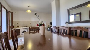 Un coin repas avec une grande table en bois, des chaises, un vase de fleurs et une cuisine en arrière-plan.