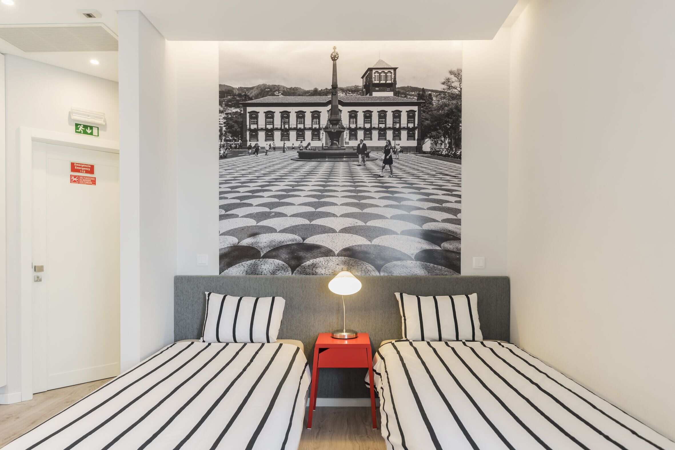 A hotel room with two beds, a nightstand, a lamp, and a large mural of a plaza.