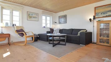 En hyggelig stue med trægulv, en grå sofa, et rundt sofabord, en gyngestol og et skab med glaslåger.