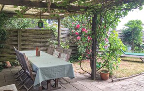 Een overdekt terras met een tafel en stoelen, omringd door een houten omheining en een bloeiende klimplant.