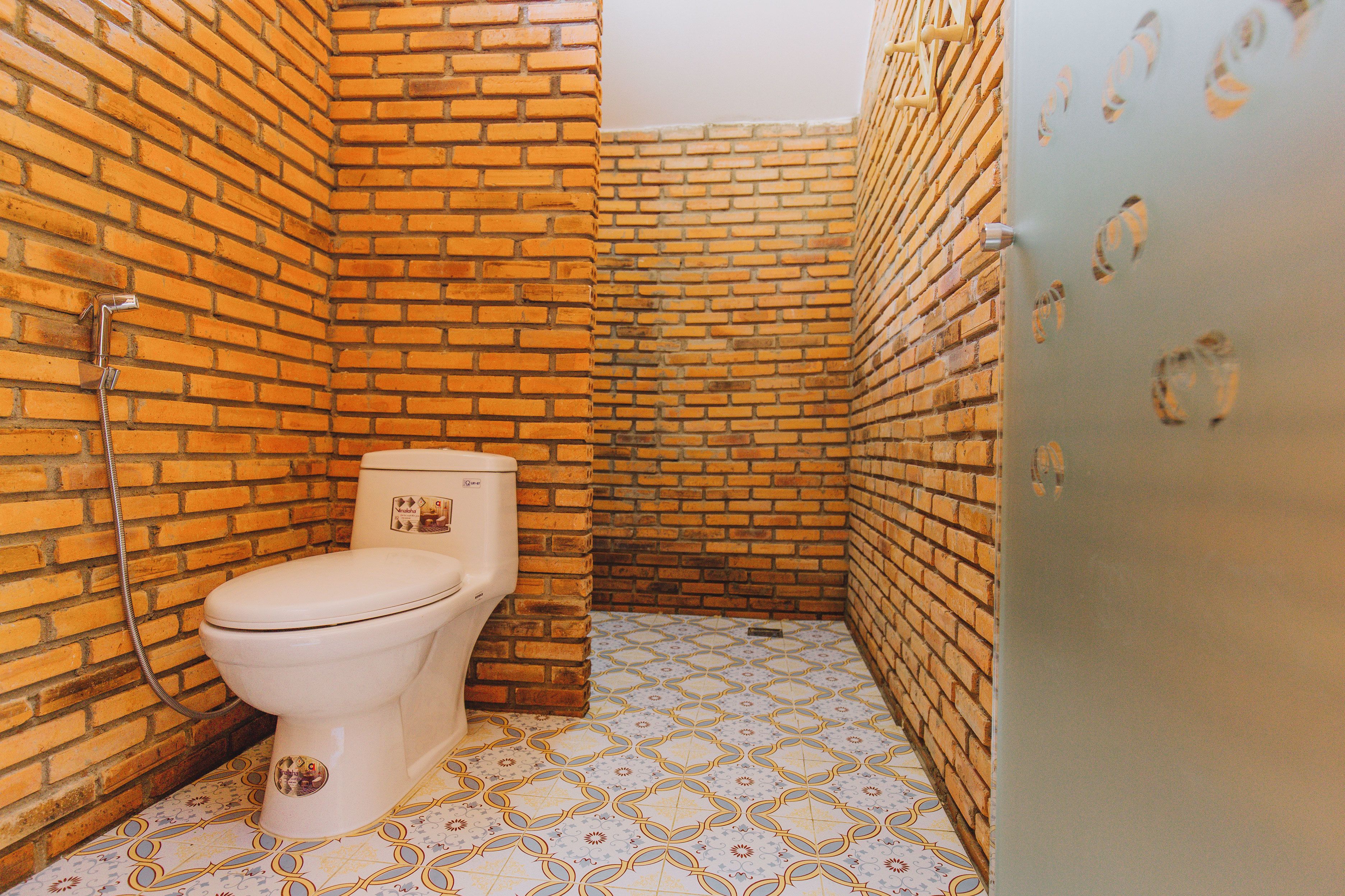 Ein kleines Badezimmer mit Toilette, Dusche und Waschbecken. Die Wände sind aus Ziegeln, und der Boden ist mit einem gemusterten Fliesenbelag versehen.