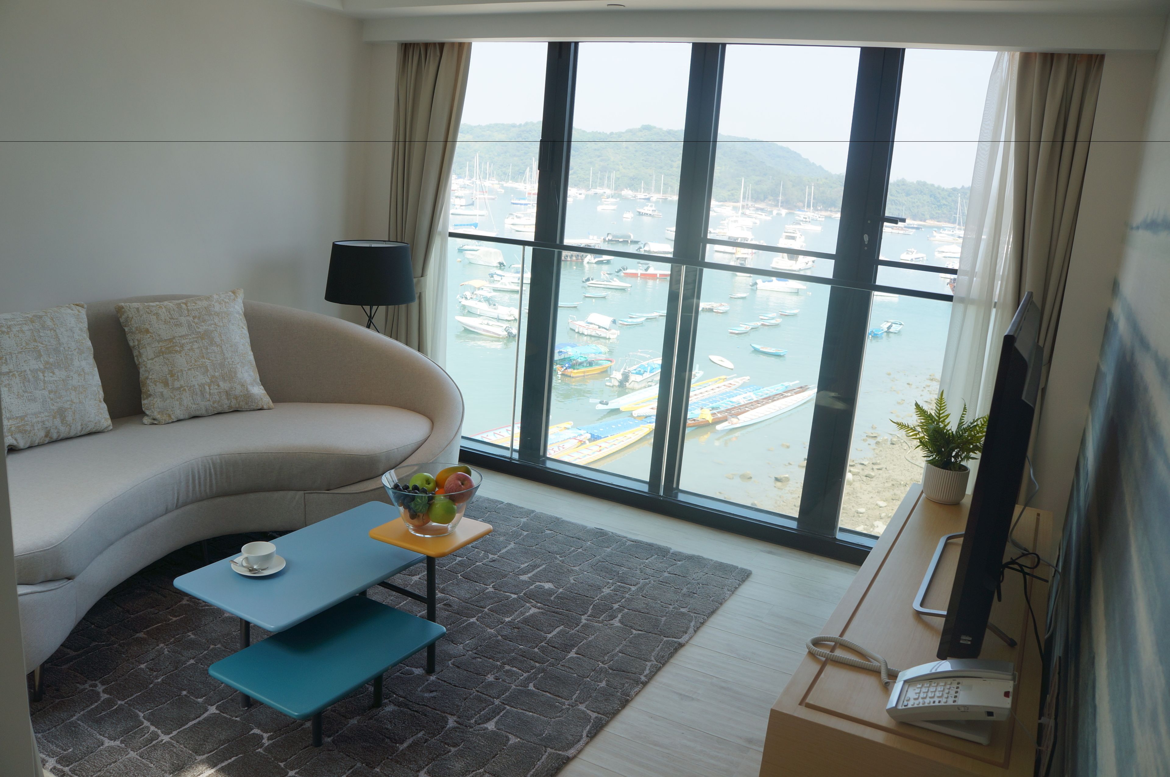 A living room with a sofa, a small table with a bowl of fruit, a TV, and a view of a marina through large windows.