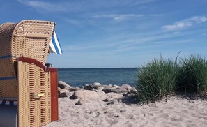 Eine Strandansicht mit einem Korbstuhl und einem gestreiften Handtuch, einer felsigen Küstenlinie und hohem Gras.