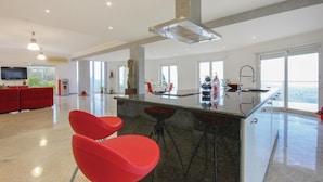 A modern kitchen with a large island, red bar stools, and a high ceiling with a central ventilation system.