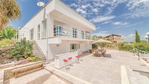 A modern two-story house with a balcony, outdoor seating, and a staircase leading to the entrance.