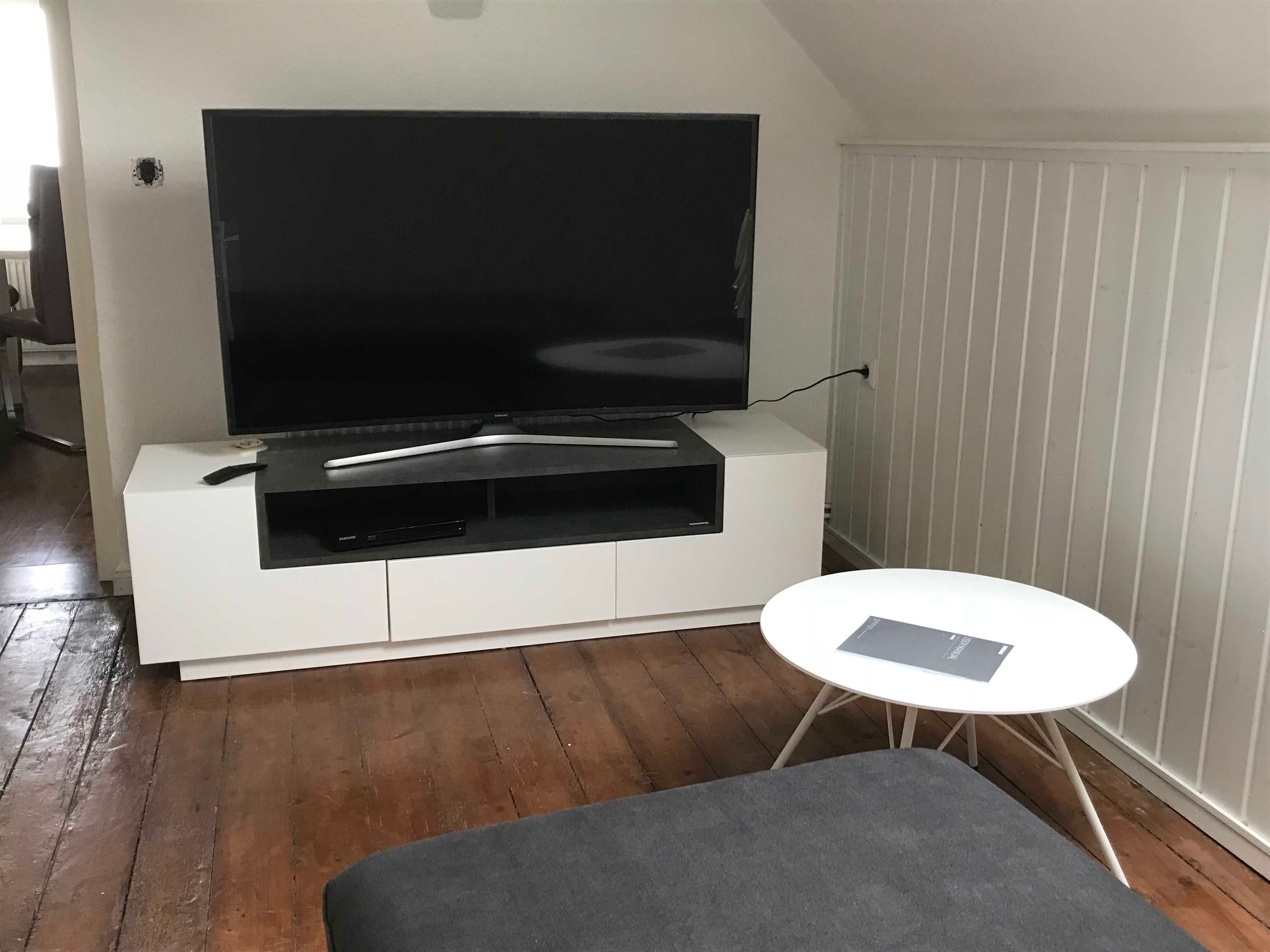 A living room with a flat-screen TV on a white entertainment unit, a small round table, and a grey sofa.