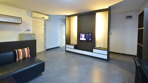 A modern hotel room with a dark-colored sofa, a flat-screen TV mounted on the wall, a built-in bench, and a small refrigerator. - LOFT Salaya (Phutthamonthon)