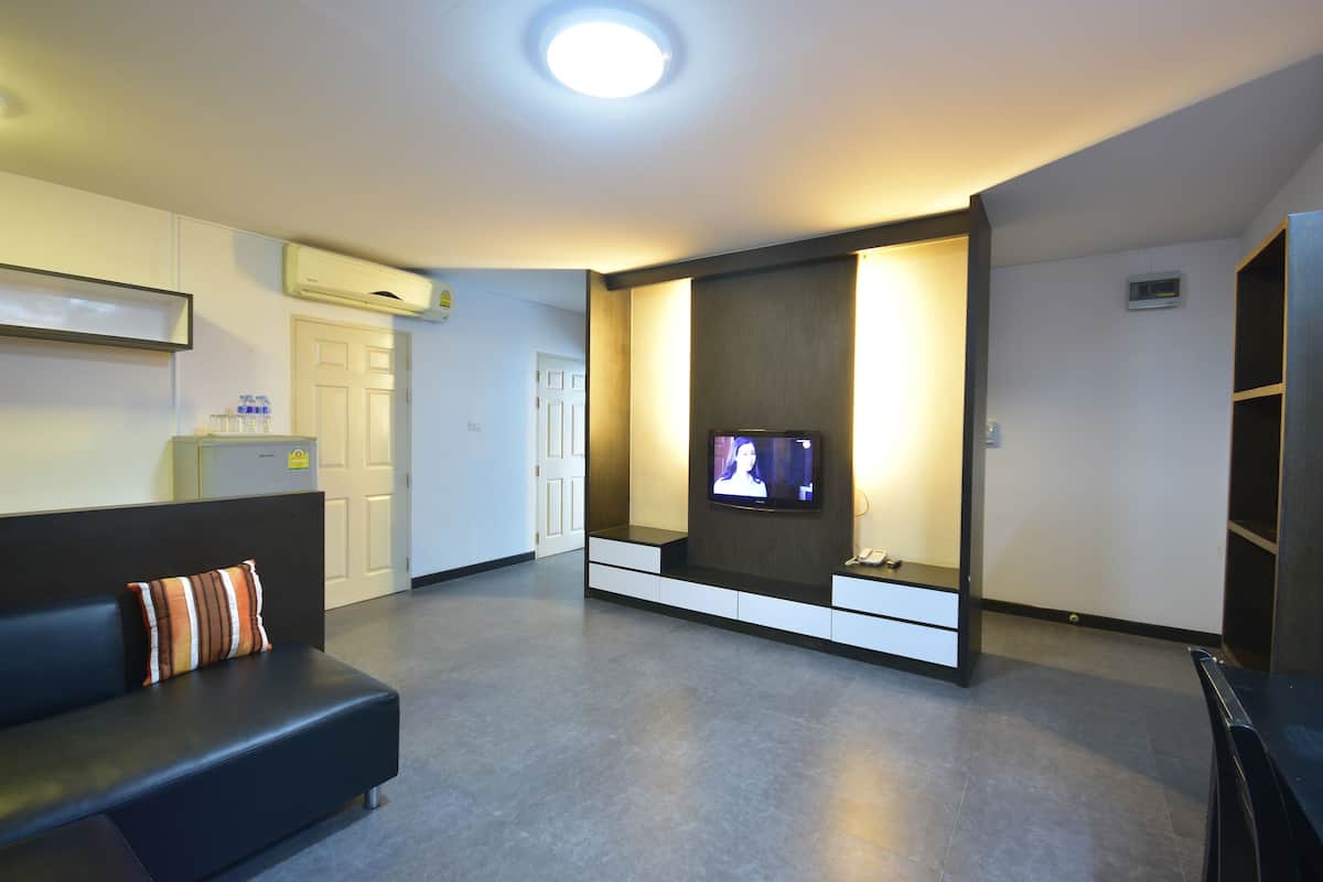 A modern hotel room with a dark-colored sofa, a flat-screen TV mounted on the wall, a built-in bench, and a small refrigerator.