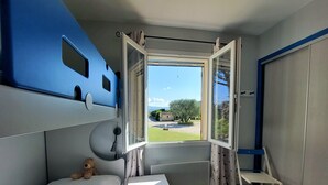 A bedroom with a bed, a desk, a chair, a window with curtains, and a view of the outdoors.