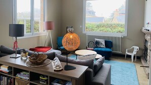 A modern living room with a sofa, a coffee table, a lamp, and a view of greenery through large windows. - La Baïta spacious house 200m from the beach (Trégastel)