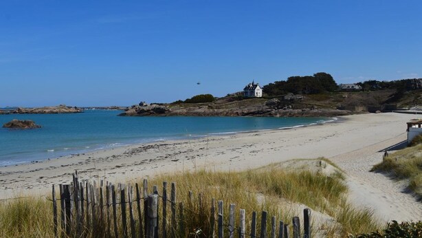 A beach with a white building on a rocky outcrop, clear blue water, and a wooden fence in the foreground. - La Baïta spacious house 200m from the beach (Trégastel)
