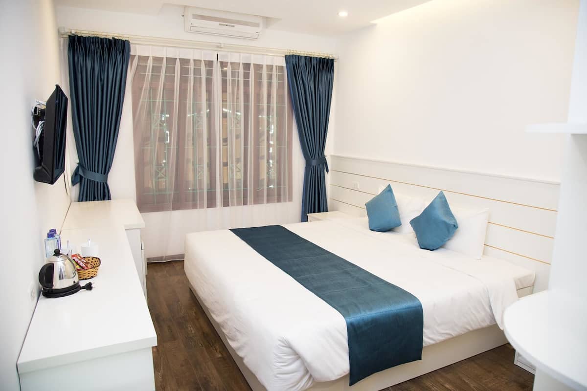 Quarto de hotel com cama, mesa de cabeceira, luminária, cesta e janela com cortinas.