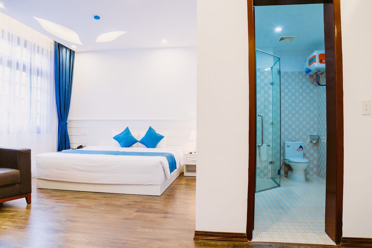 Quarto de hotel com cama, poltrona, mesa de cabeceira e banheiro com chuveiro e vaso sanitário.