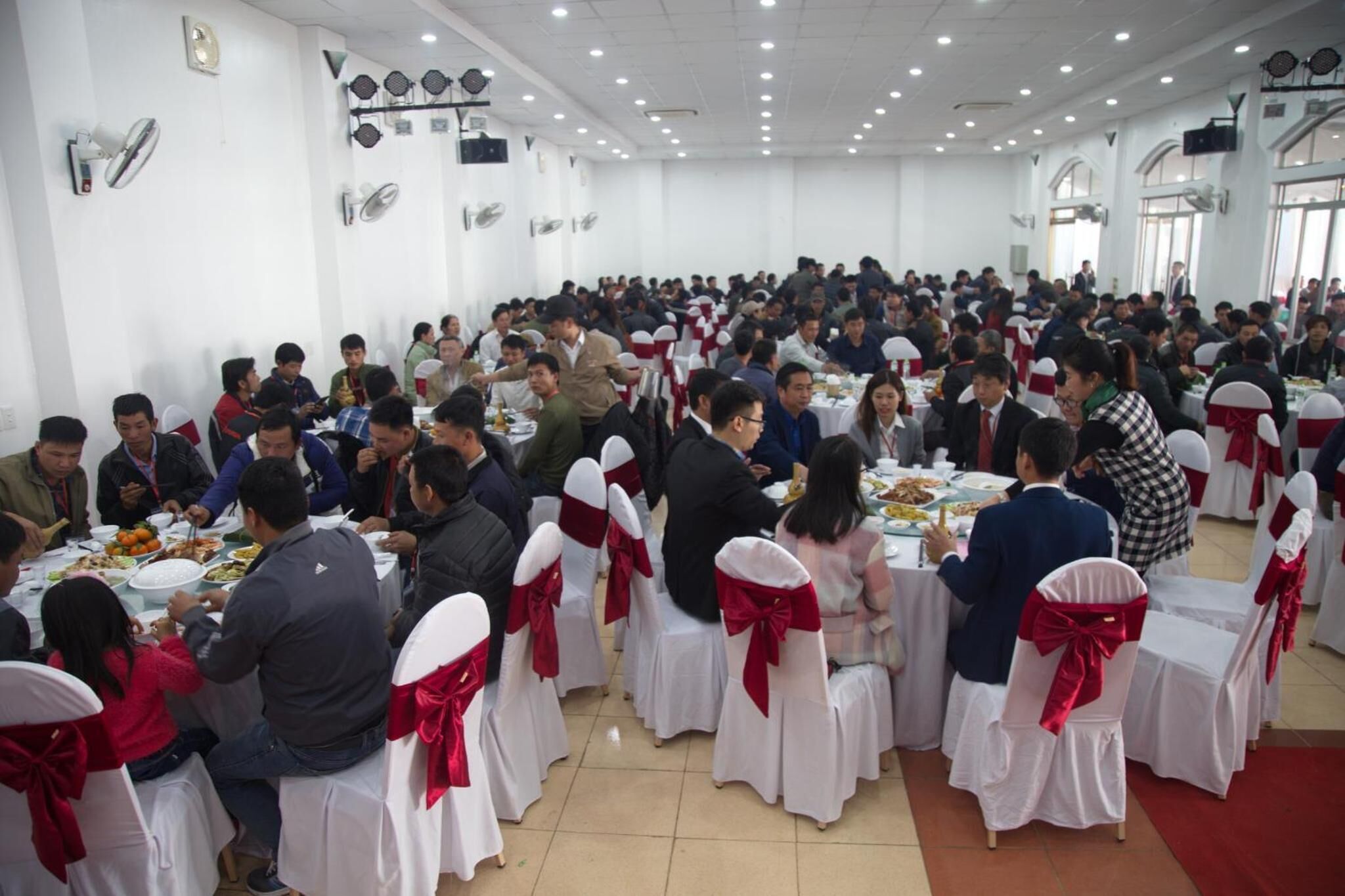 Un vaste espace de restauration intérieur, avec des tables rondes et des chaises blanches, dont certaines sont ornées de rubans rouges.