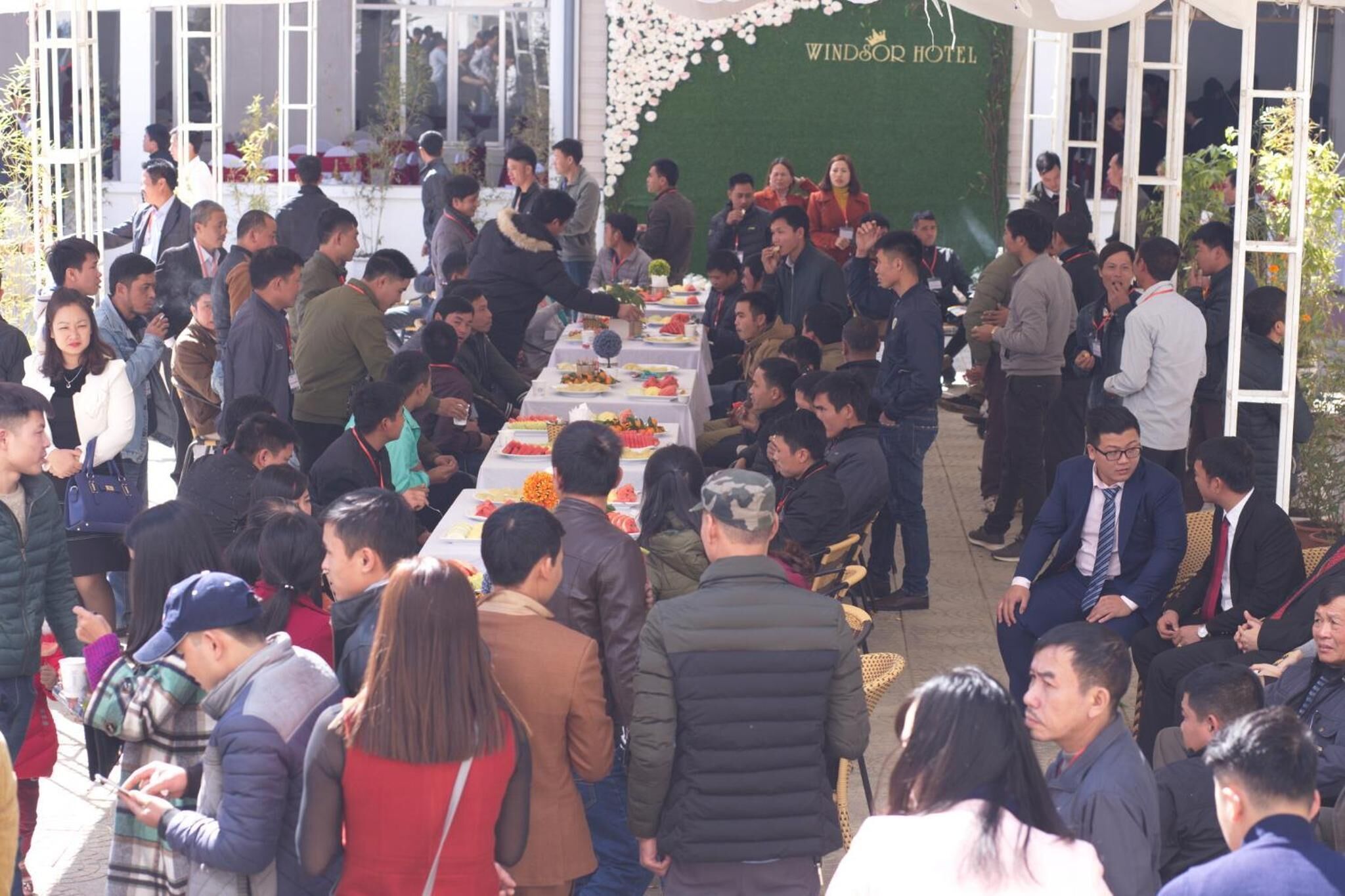 A crowded outdoor dining area with tables set for a meal, surrounded by a green wall and a sign reading "WIND&OR HOTEL".