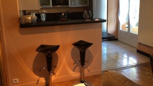 Cozinha compacta com micro-ondas, chaleira e máquina de café no balcão. Há dois bancos em frente ao balcão.