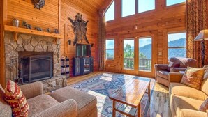 L’intérieur d’une cabane en rondins, avec une cheminée en pierre, des meubles en bois et de grandes fenêtres offrant une vue sur les montagnes.