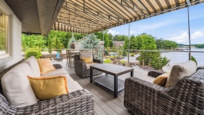 A covered outdoor seating area with wicker furniture, a table, and a view of a lake and greenery. - "Bayshore Breeze" - Lakefront 7BR, Private Pool, Hot Tub, Putting Green, Views (Hot Springs)