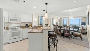 A modern kitchen with white cabinetry, a granite countertop, and a central island with bar stools. There are ceiling lights and a view of the ocean through large windows. - Spacious Oceanfront Condo, Jacuzzi Tub, Lazy River | Mar Vista Grande - 1206 (North Myrtle Beach)