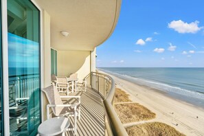 A balcony with a view of the beach, a table, and chairs.