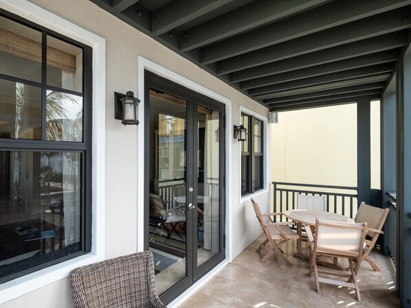 A covered outdoor area with a table and chairs, a glass door leading to an interior space, and a balcony with a railing.