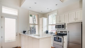 A kitchen with white cabinetry, a central island, stainless steel appliances, and a view through the windows. - French Quarter Escape – Walk to City Market, Rainbow Row & More (Charleston)