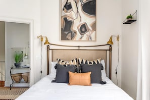 A neatly arranged bedroom with a bed, bedside lamps, a wall art piece, and a small shelf with a plant.