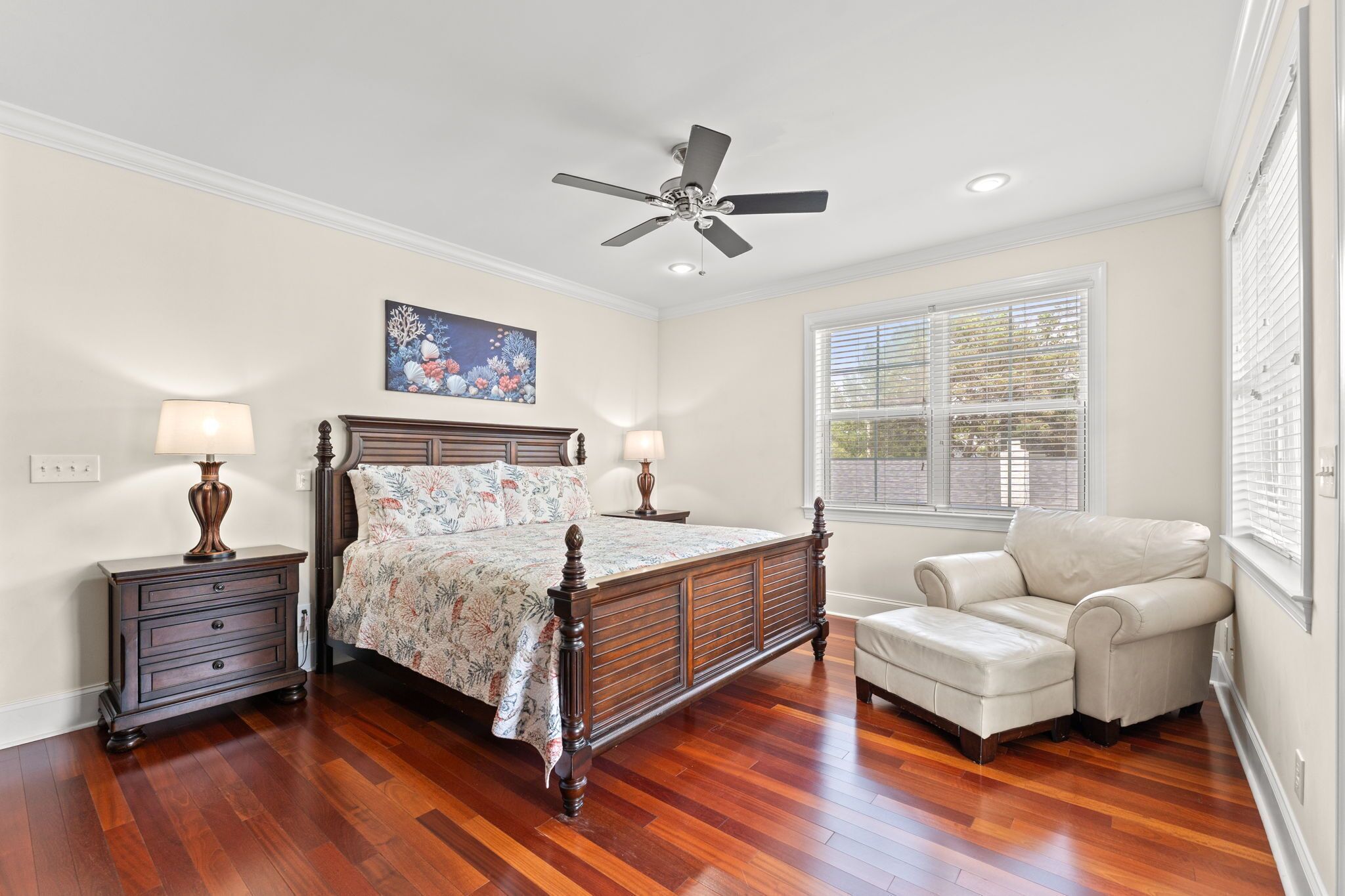 A bedroom with a wooden bed, two bedside tables with lamps, a ceiling fan, a window with blinds, and a sofa.