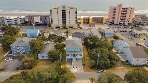 Vue aérienne d’une ville côtière avec des hôtels en bord de mer, un bâtiment central à la façade bleue et blanche, et un parking.