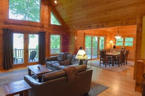 A wooden cabin interior with a living area, dining table, and large windows.