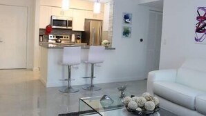 Una sala moderna con un sofá blanco, una mesa de centro de vidrio y una zona de cocina con microondas, estufa y dos taburetes de bar.
