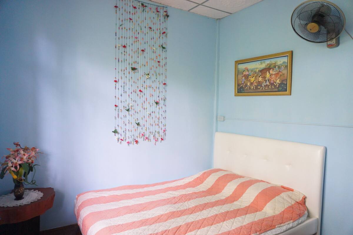 Een slaapkamer met een bed, een wandventilator, een lijst met een afbeelding en een bloemen gordijn.