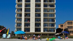 Un hotel de varios pisos con balcones en cada planta, situado cerca de una playa con sillas y sombrillas.
