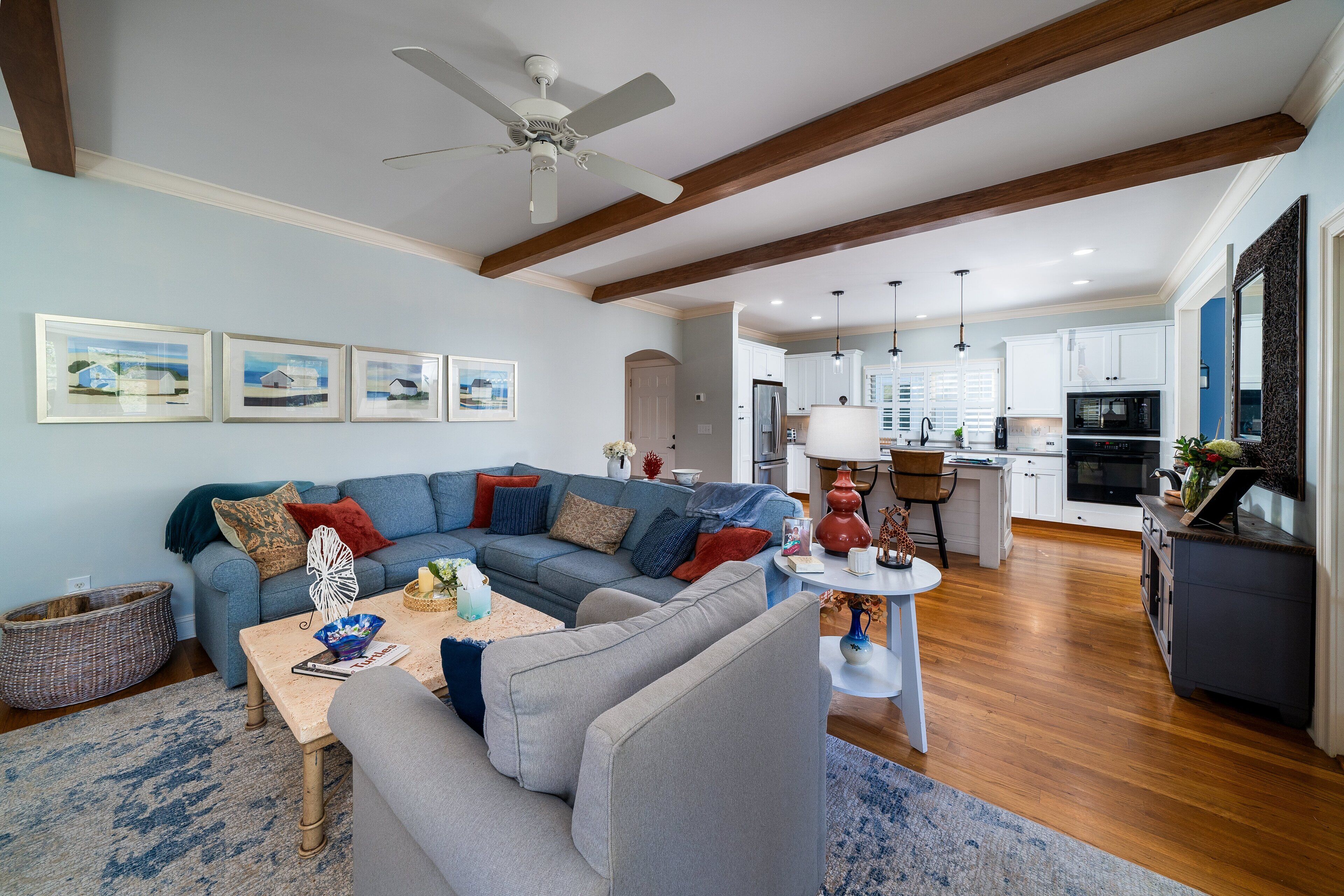 A modern living room with a sectional sofa, a coffee table, and a dining area with a small table and chairs. There is a kitchen in the background with white cabinets and stainless steel appliances.