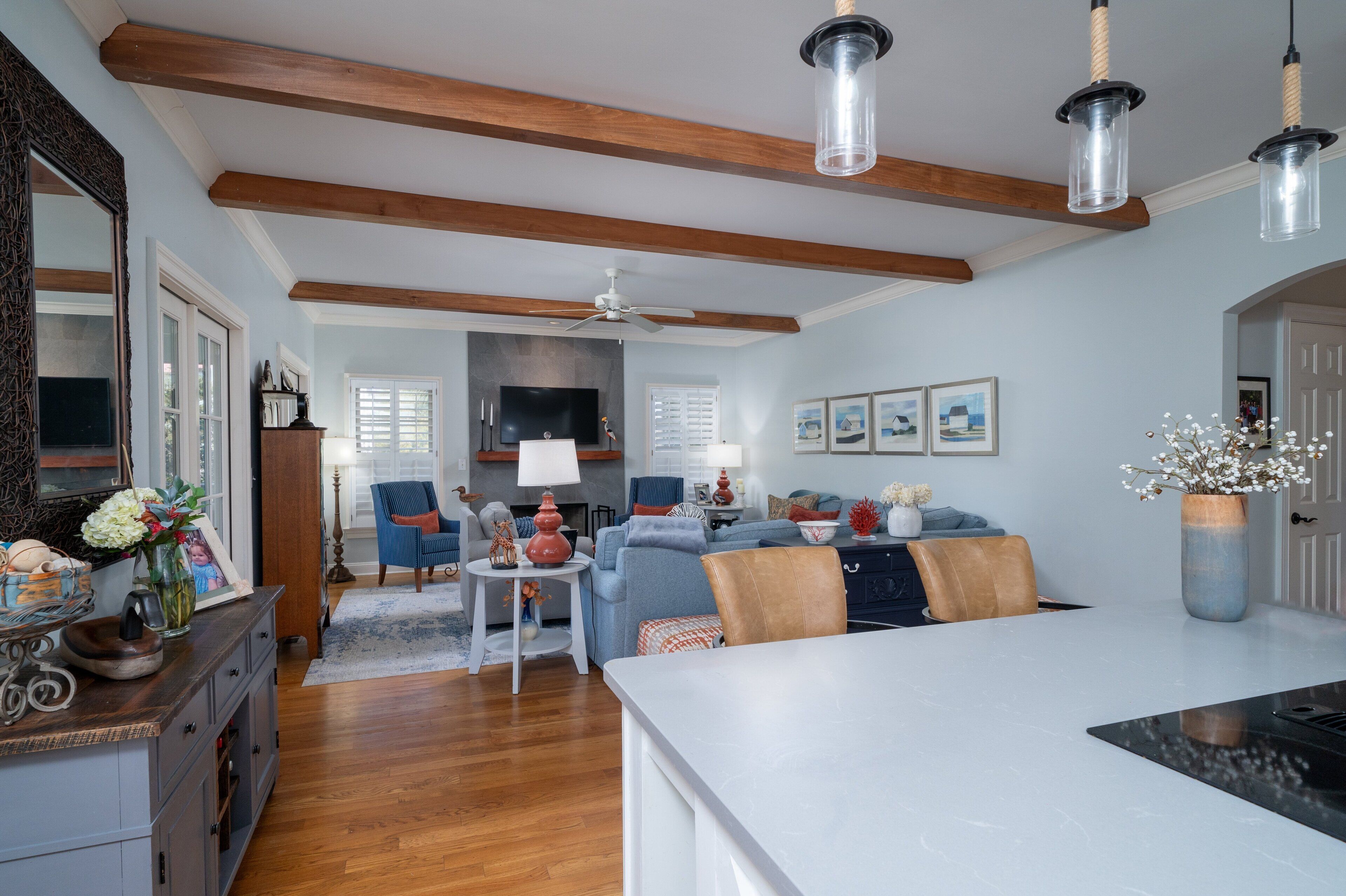 Un salon moderne doté d’un grand îlot central blanc, de poutres en bois au plafond, d’un ventilateur de plafond et d’un téléviseur fixé au mur.