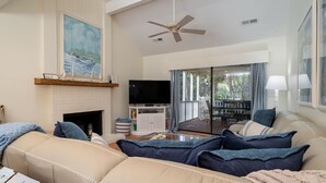 Amplia sala de estar con un sofá color beige, televisor de pantalla plana, chimenea y vistas al exterior a través de grandes ventanales.