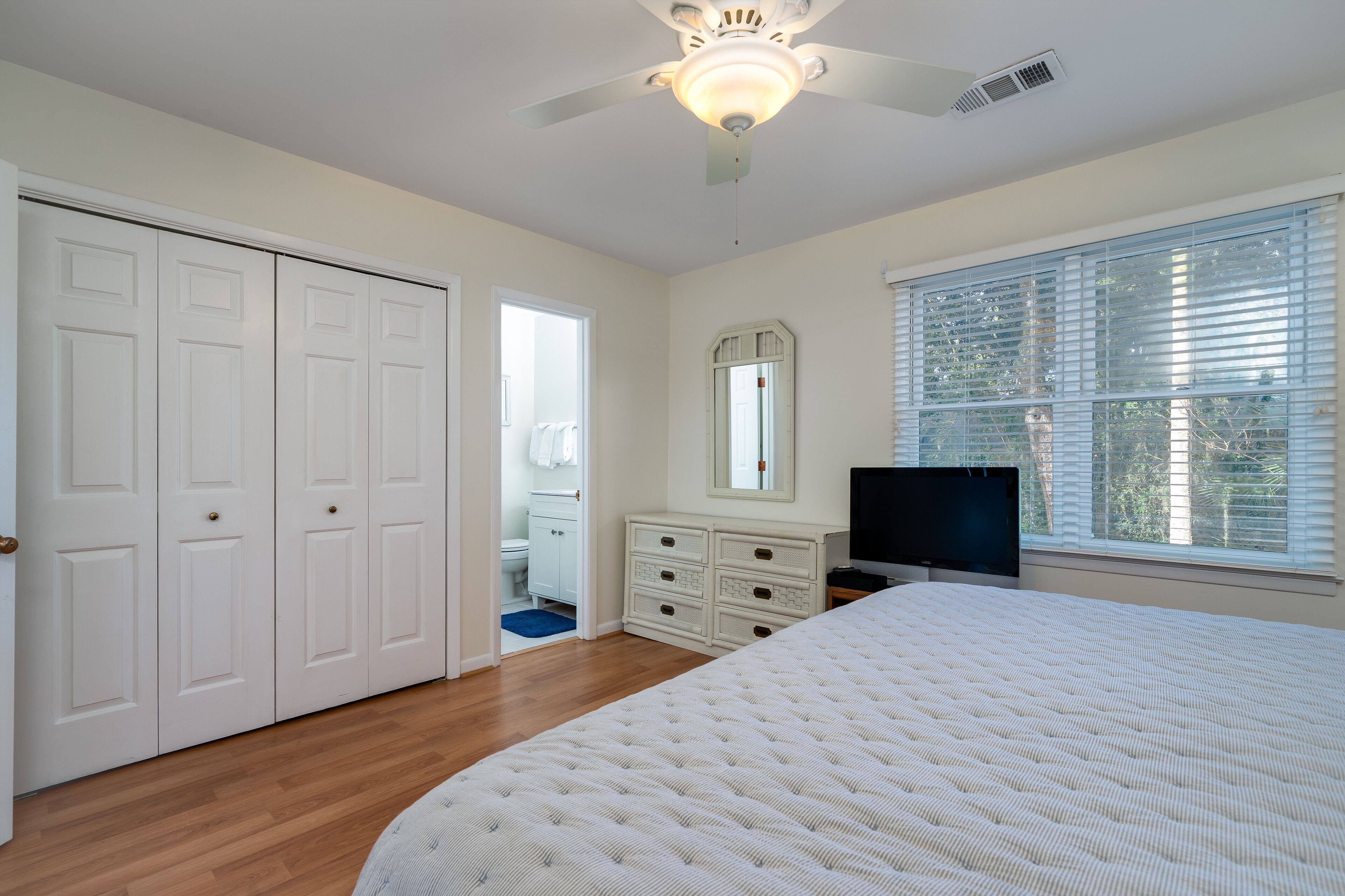 A bedroom with a large bed, a ceiling fan, a dresser, a TV, and a window with blinds.