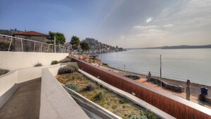 Une promenade en bord de mer, avec un chemin en béton, des bancs en bois et une vue sur l’eau et les bâtiments situés au loin.