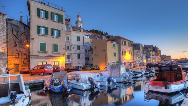 Un port de plaisance avec des bateaux amarrés, une rue bordée d’immeubles et un ciel dégagé.