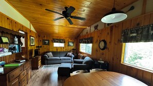 A cozy cabin interior with a wooden ceiling, a ceiling fan, a large window, a sofa, a dining table, and a clock on the wall. - Lake Front Cabin with free WiFi - Adirondacks High Peaks Region - Lake Placid (Elizabethtown)