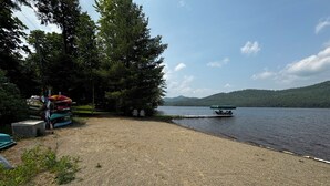 A gravel beach with kayaks and a paddleboard, a covered dock, and a rowboat on a calm lake with a forested hill in the background. - Lake Front Cabin with free WiFi - Adirondacks High Peaks Region - Lake Placid (Elizabethtown)