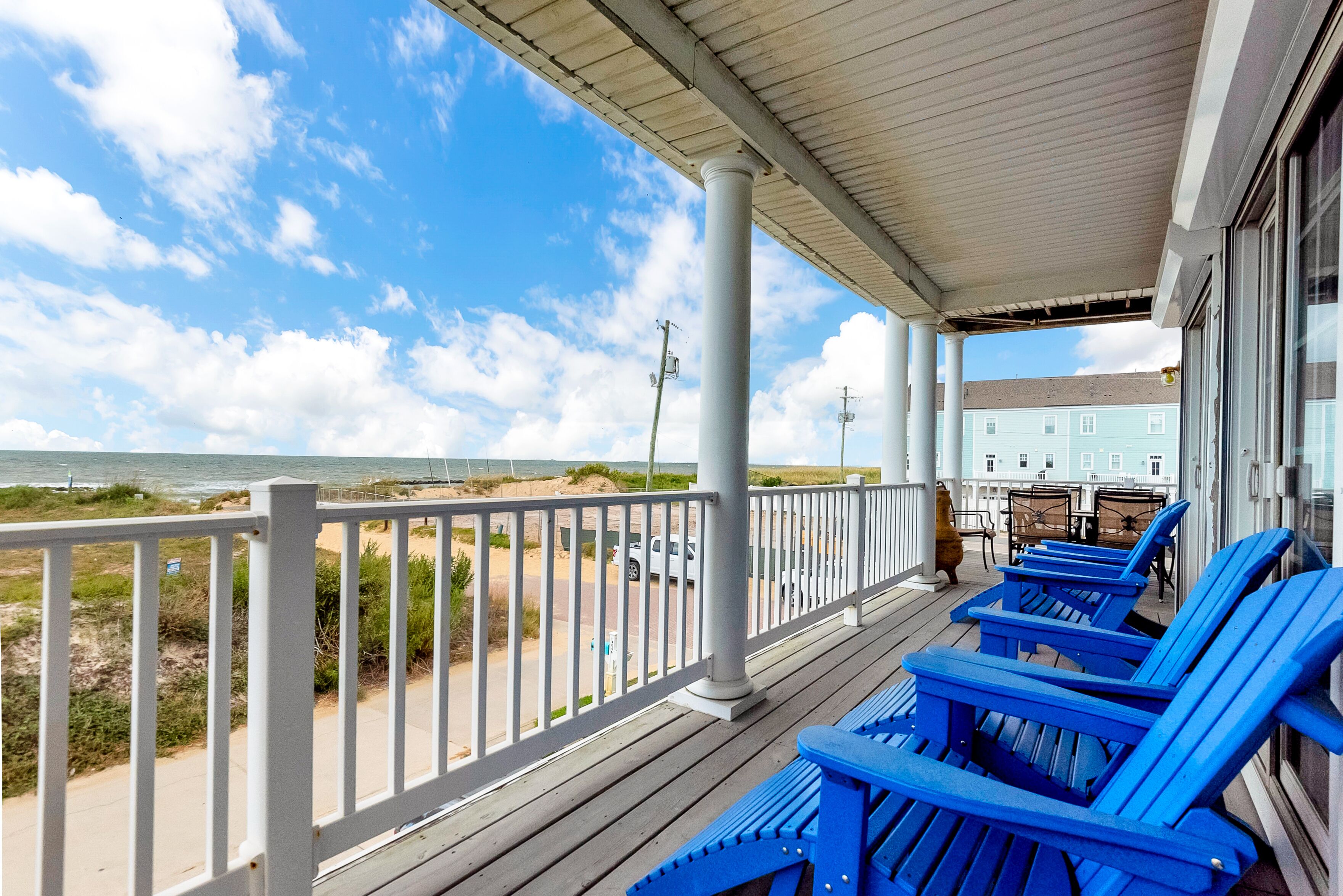 A covered outdoor area with a view of the beach, featuring a railing, blue chairs, and a building in the background.