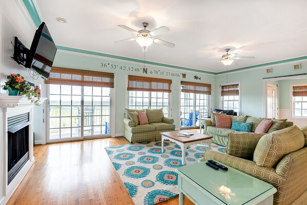 A living room with a fireplace, two ceiling fans, a television, and a large window with blinds.