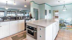 A modern kitchen with white cabinets, stainless steel appliances, and a large island. There are pendant lights, a dining area with a table and chairs, and a colorful rug. - 3-Story Oceanfront • Breathtaking Views • Dogs OK (Norfolk)