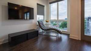 Un salon moderne doté d’un grand téléviseur à écran plat fixé au mur, d’un fauteuil inclinable en cuir, d’un meuble en bois foncé et d’une grande fenêtre offrant une vue sur la ville.