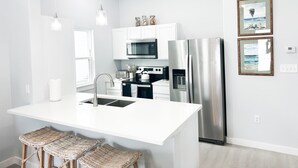 Une cuisine moderne dotée d’un îlot central, d’appareils électroménagers en acier inoxydable, de meubles blancs et de tabourets de bar en osier.