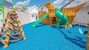 Une aire de jeux avec un toboggan en bois, une structure d’escalade faite de cordes et un espace de jeux sur le thème des bateaux.